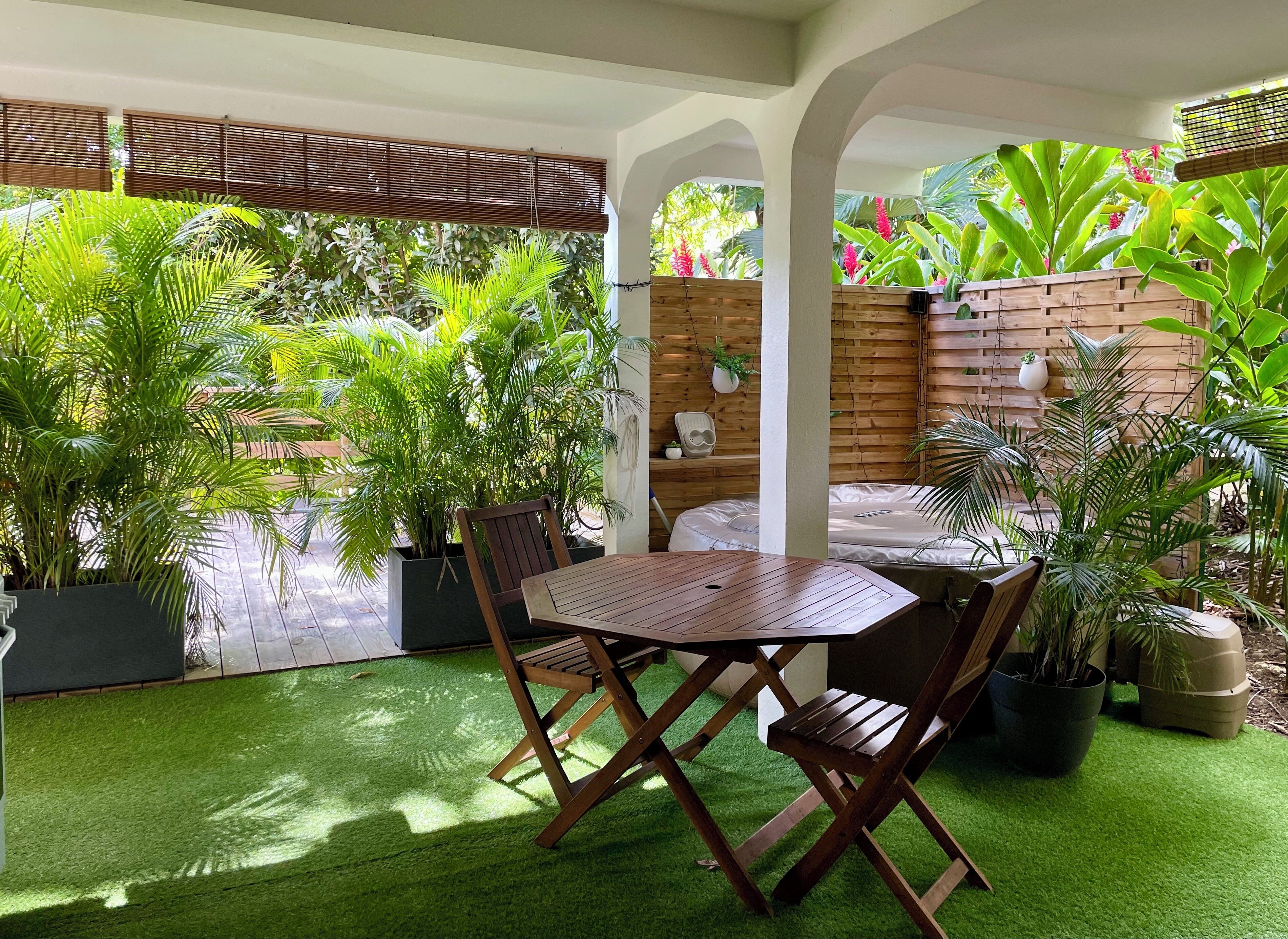 Un patio al aire libre con una mesa y sillas de madera, rodeado de plantas verdes exuberantes y una cerca de madera.