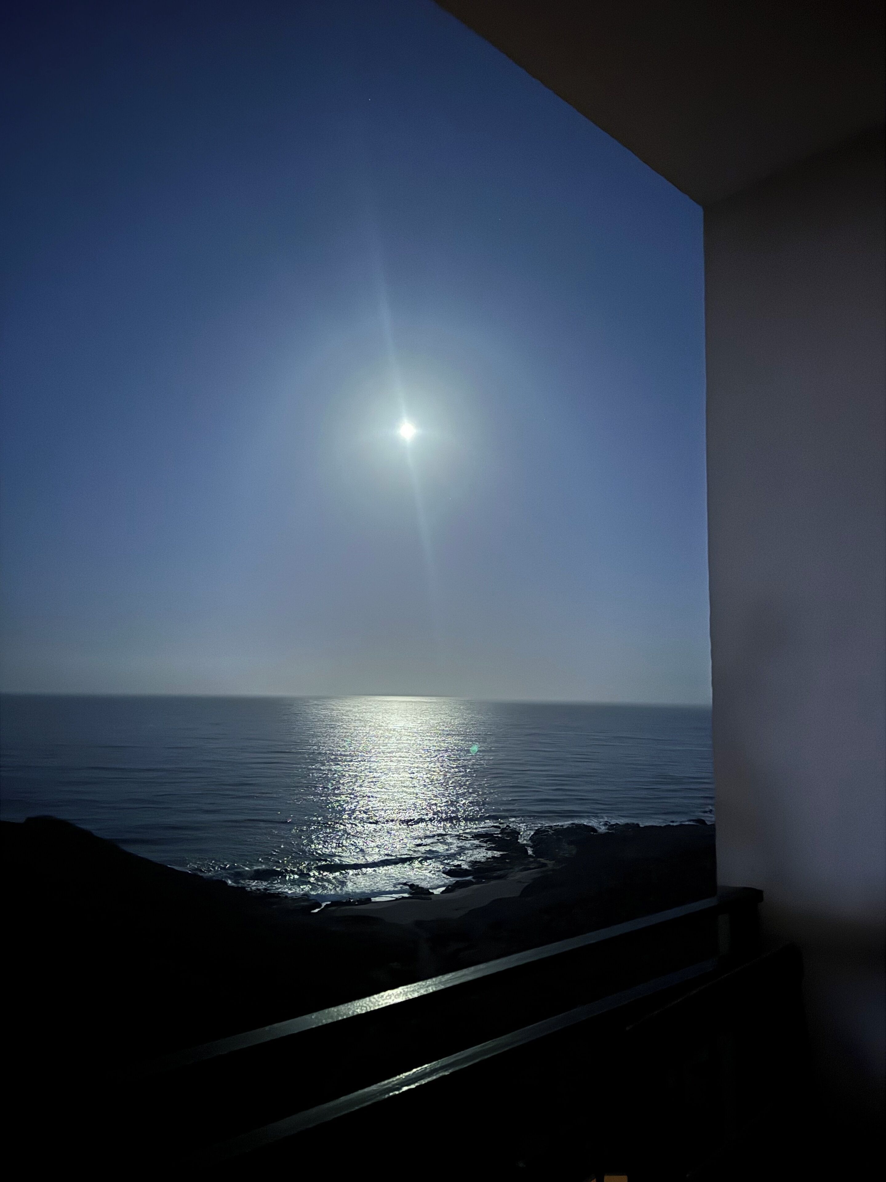 A view of the ocean with the sun reflecting off the water, a railing, and a building in the foreground.