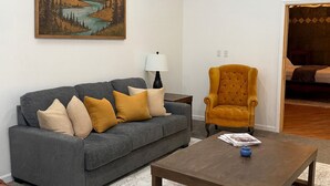 Uma sala de estar com um sofá cinza, um banquinho de couro marrom, uma mesa de centro de madeira e uma poltrona amarela mostarda. Um quadro de uma paisagem montanhosa está pendurado na parede.