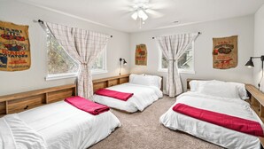A room with three single beds, white bedding, and red throw pillows. There are posters on the walls, a ceiling fan, and a window with curtains.