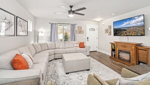 A living room with a sectional sofa, ottoman, fireplace, flat-screen TV, ceiling fan, and artwork on the walls.
