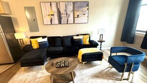 A modern living room with a sectional sofa, a round coffee table, and a blue armchair. There are abstract paintings on the wall and a window with curtains.