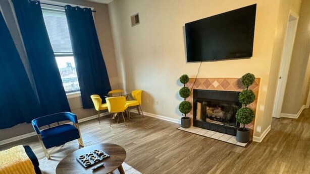 A living room with a fireplace, a flat-screen TV mounted on the wall, and a dining area with yellow chairs.
