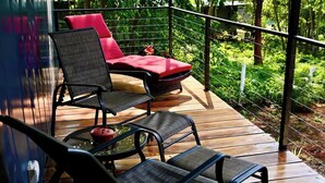 Une terrasse en bois avec deux chaises noires et un coussin rouge, une petite table avec un saladier, et une balustrade donnant sur un jardin luxuriant et verdoyant.
