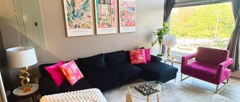 Ein modernes Wohnzimmer mit einer schwarzen Couch, einem weißen Pouf, einem Glastisch und einem rosa Sessel. An der Wand hängen drei gerahmte Kunstwerke und es gibt ein großes Fenster mit Vorhängen.