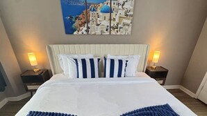 Un lit avec une literie blanche et des oreillers bleus, une couverture bleue tricotée et un tableau représentant un paysage côtier, accroché au-dessus du lit.