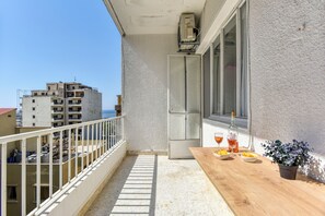 Een balkon met een houten tafel, twee glazen, een potplant en een raam met uitzicht op gebouwen.