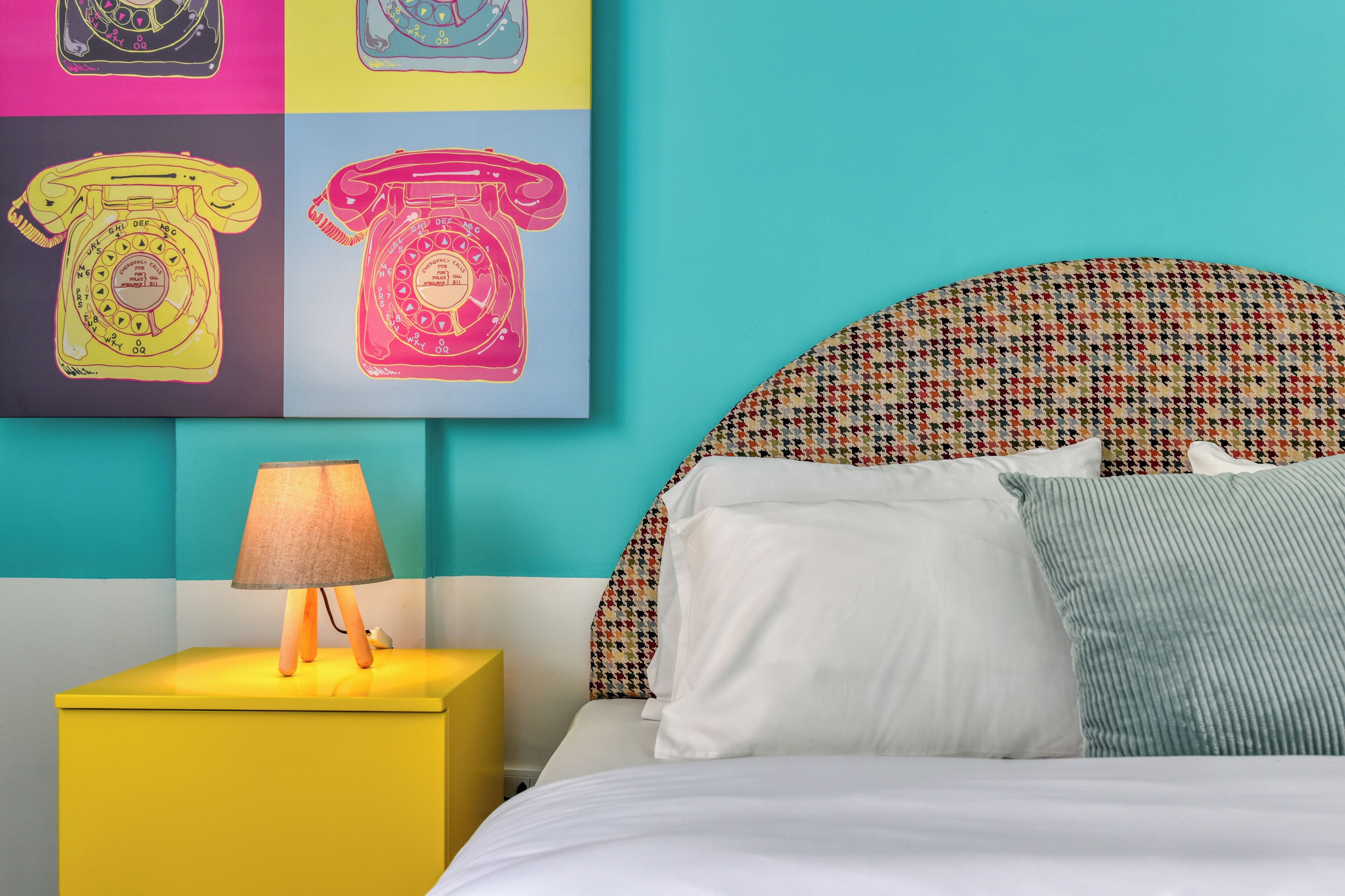 A bed with white and blue pillows, a patterned headboard, and a yellow bedside table with a lamp.