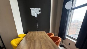 Quarto moderno com mesa de madeira, cadeiras amarelas e laranjas, parede preta com o letreiro 'Welcome to Harry Potter' e janela com vista para edifícios.