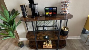 Uma estação de café em madeira, com cafeteira, caneca, bandeja de recipientes coloridos e um objeto decorativo.