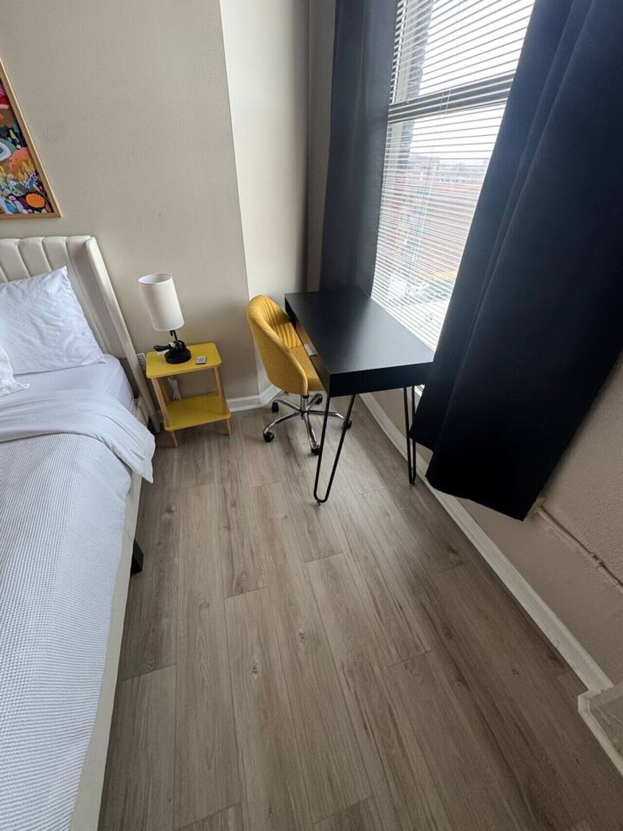 A small, neatly arranged room with a bed, a desk, a chair, and a window with blinds.
