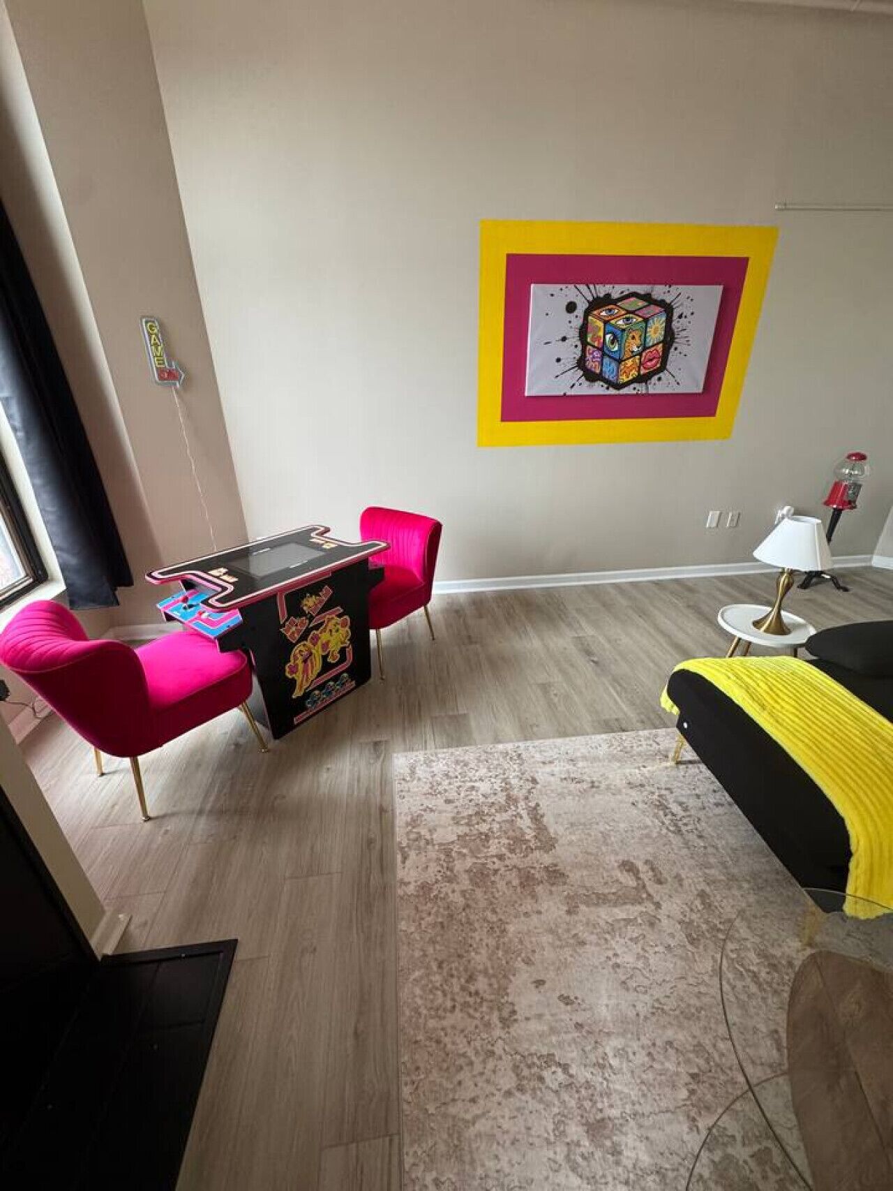 Une pièce moderne avec un plancher en bois, une table de jeu noire, une chaise rouge, un canapé jaune et noir, une lampe et une œuvre d’art colorée accrochée au mur.