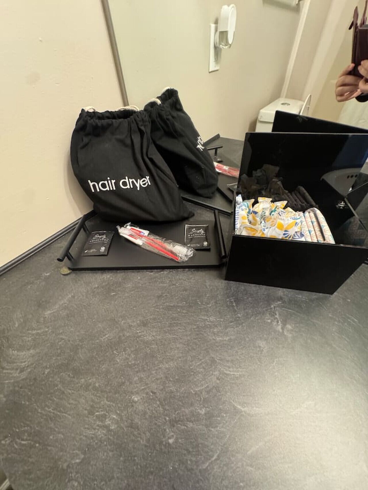 Eine schwarze Tasche für Haartrockner mit dem Aufdruck "hair dryer" auf einer schwarzen Ablage, neben einer Schachtel mit verschiedenen Haarpflegeprodukten.