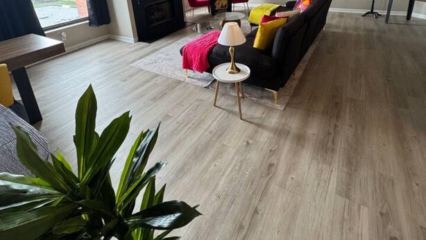 Uma sala de estar moderna com um sofá preto, uma mesinha redonda e uma televisão. Há uma janela com vista para prédios.