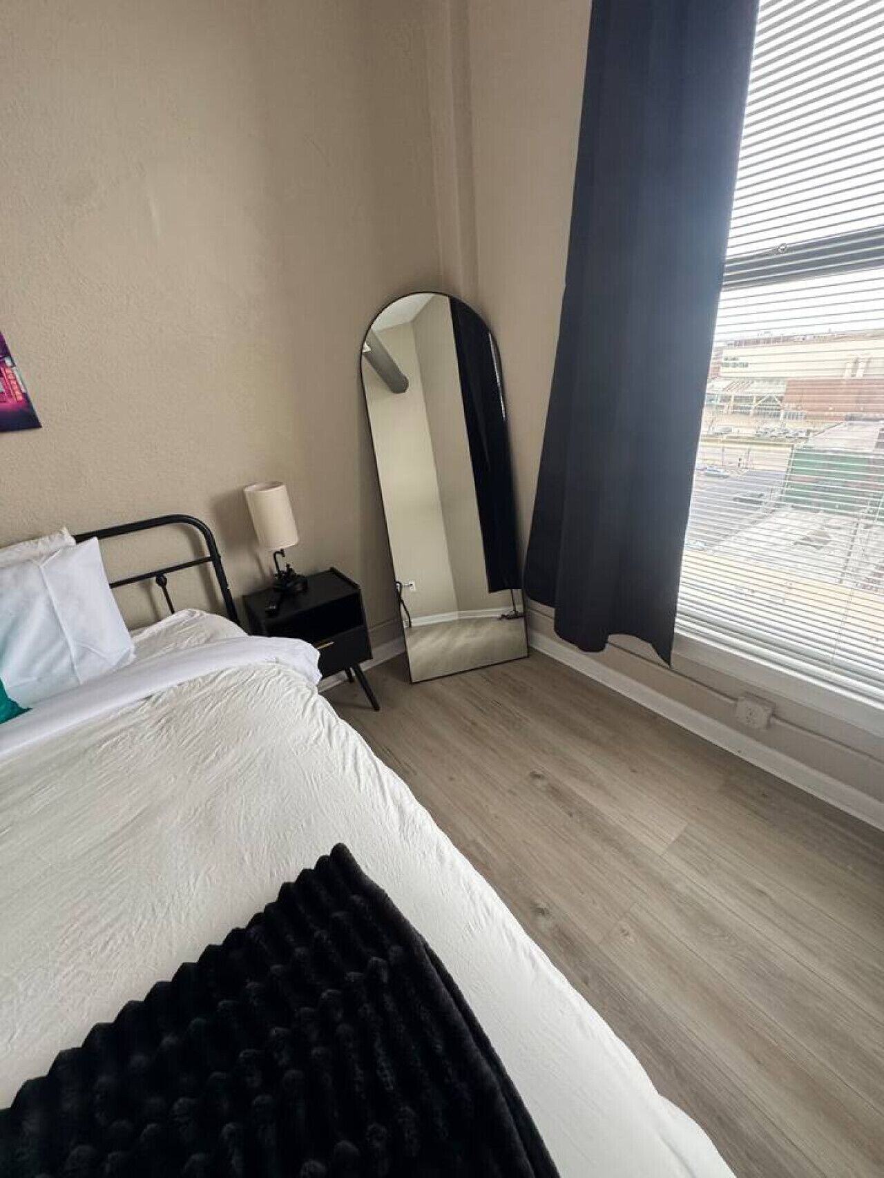 A hotel room with a bed, a nightstand, a mirror, a window with blinds, and a view of the outside.