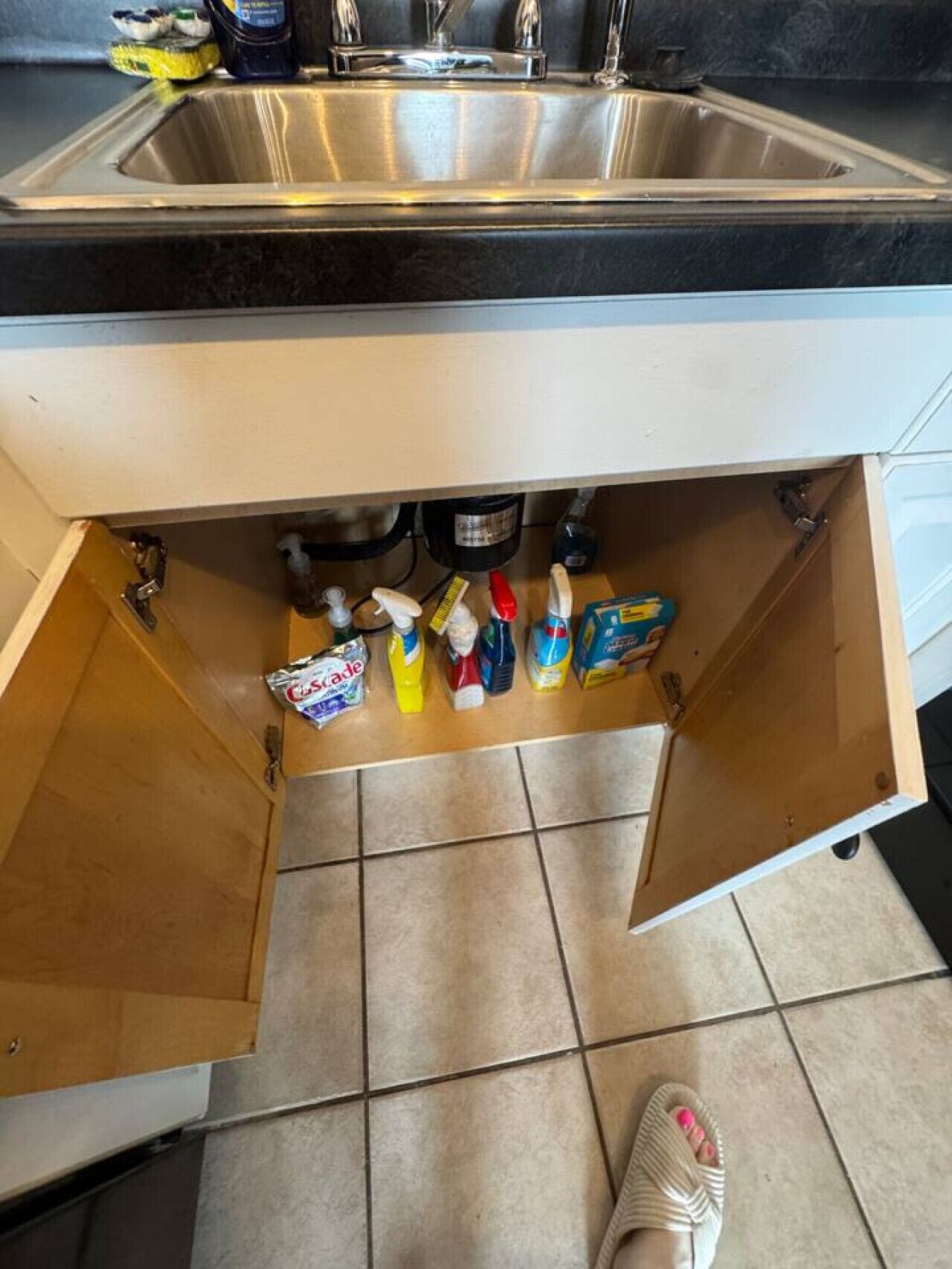 A kitchen with a stainless steel sink, a wooden cabinet with a drawer, and various cleaning supplies on the countertop.