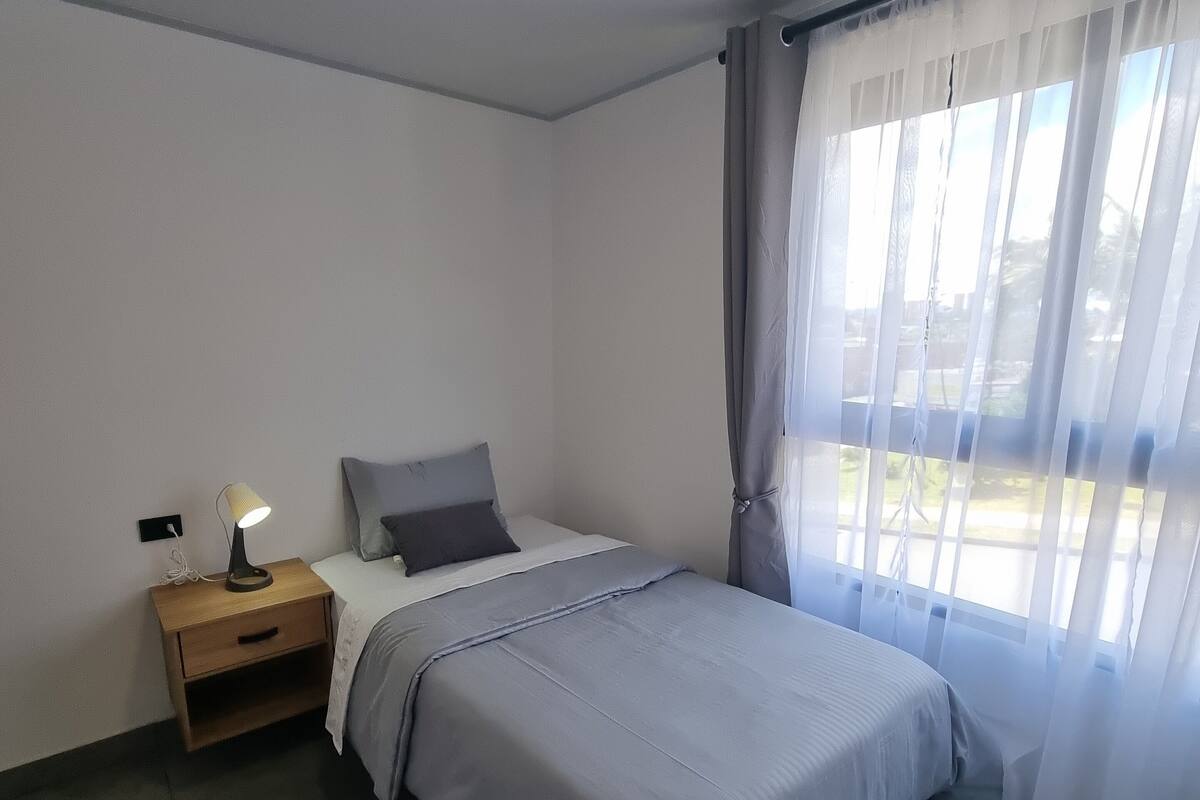 Une chambre à coucher avec un lit, une table de chevet avec une lampe, une fenêtre avec des rideaux légers et une vue sur l’extérieur.