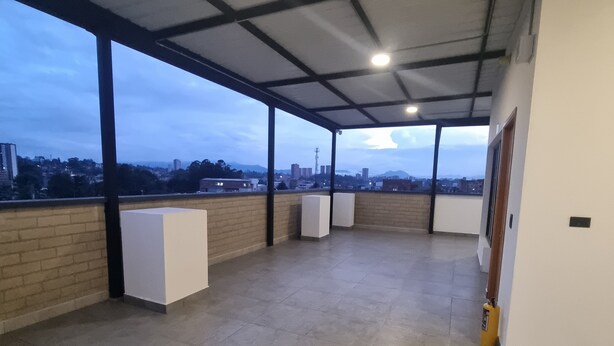 Eine offene Terrasse mit Blick auf die Stadtlandschaft, darunter Gebäude und ein Turm. Die Terrasse hat einen gefliesten Boden und eine Metall-Dachkonstruktion.