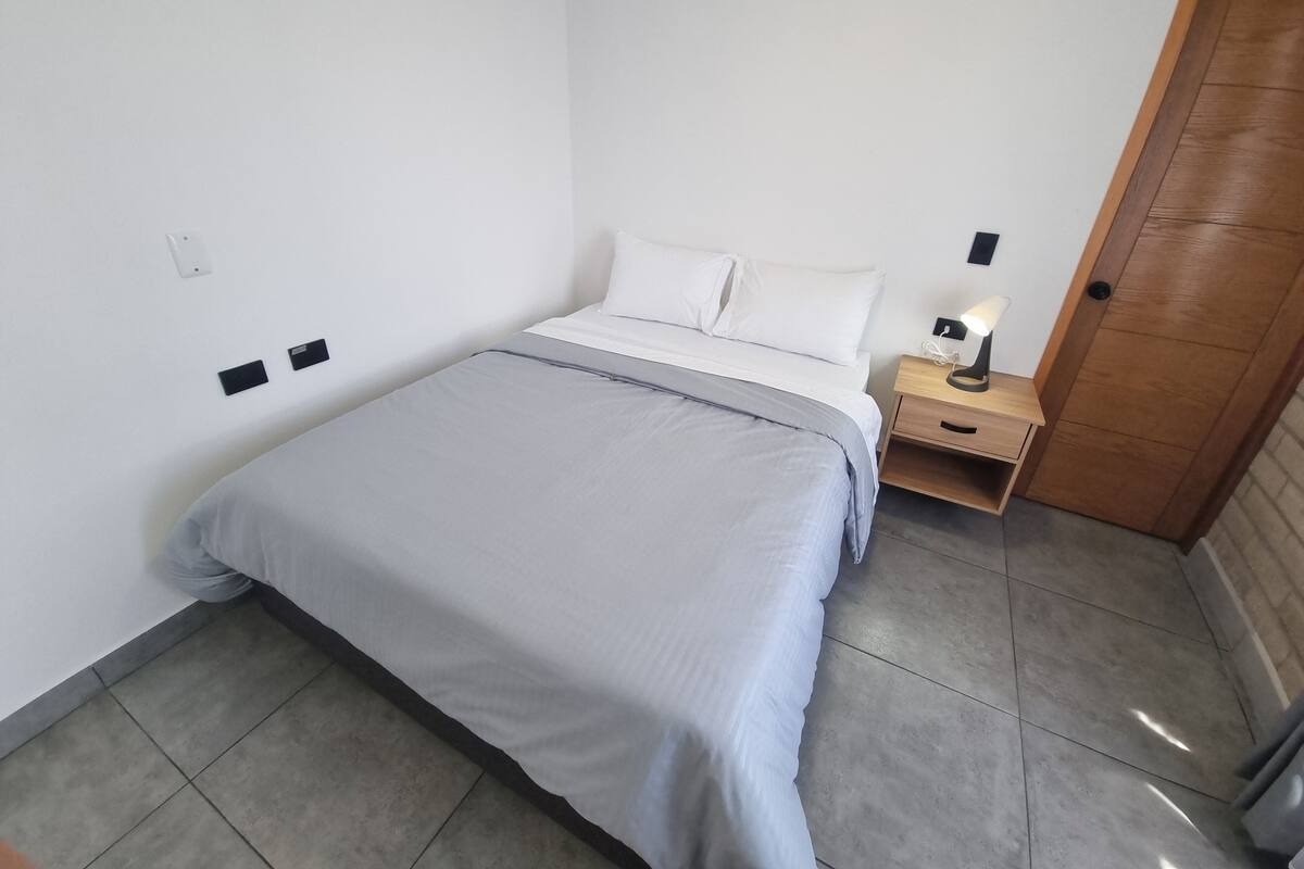 Un lit bien fait, recouvert d’une couverture grise, une table de chevet en bois avec une lampe, et une porte en bois.