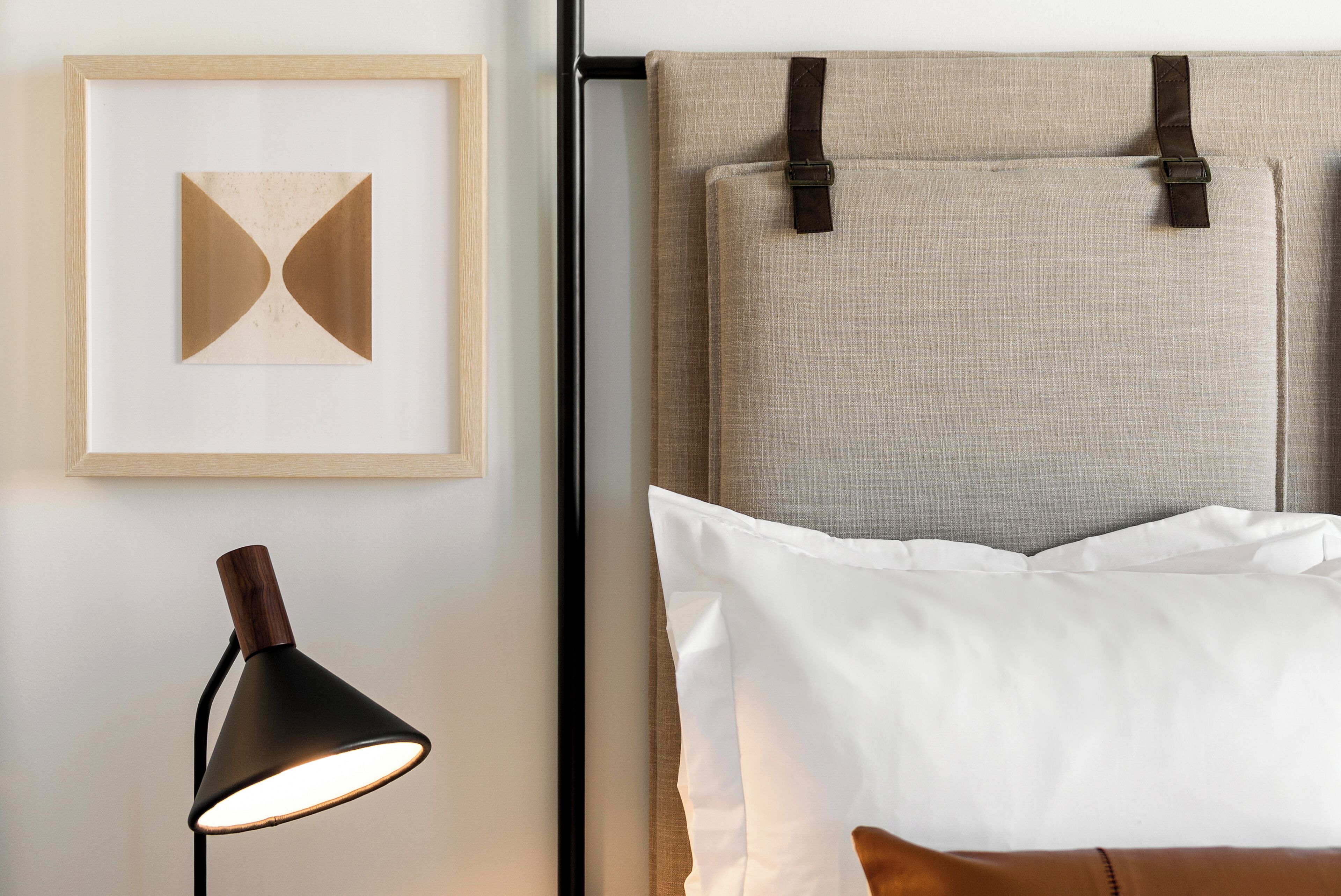 A neatly arranged bed with a headboard, a framed artwork on the wall, and a floor lamp.