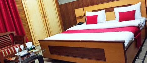 A hotel room with a wooden bed, red and white bedding, a small table with a chair, and a wardrobe.