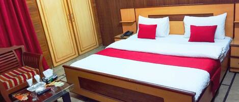 A hotel room with a large bed, a wooden headboard, a nightstand, a desk with a chair, and a wardrobe.