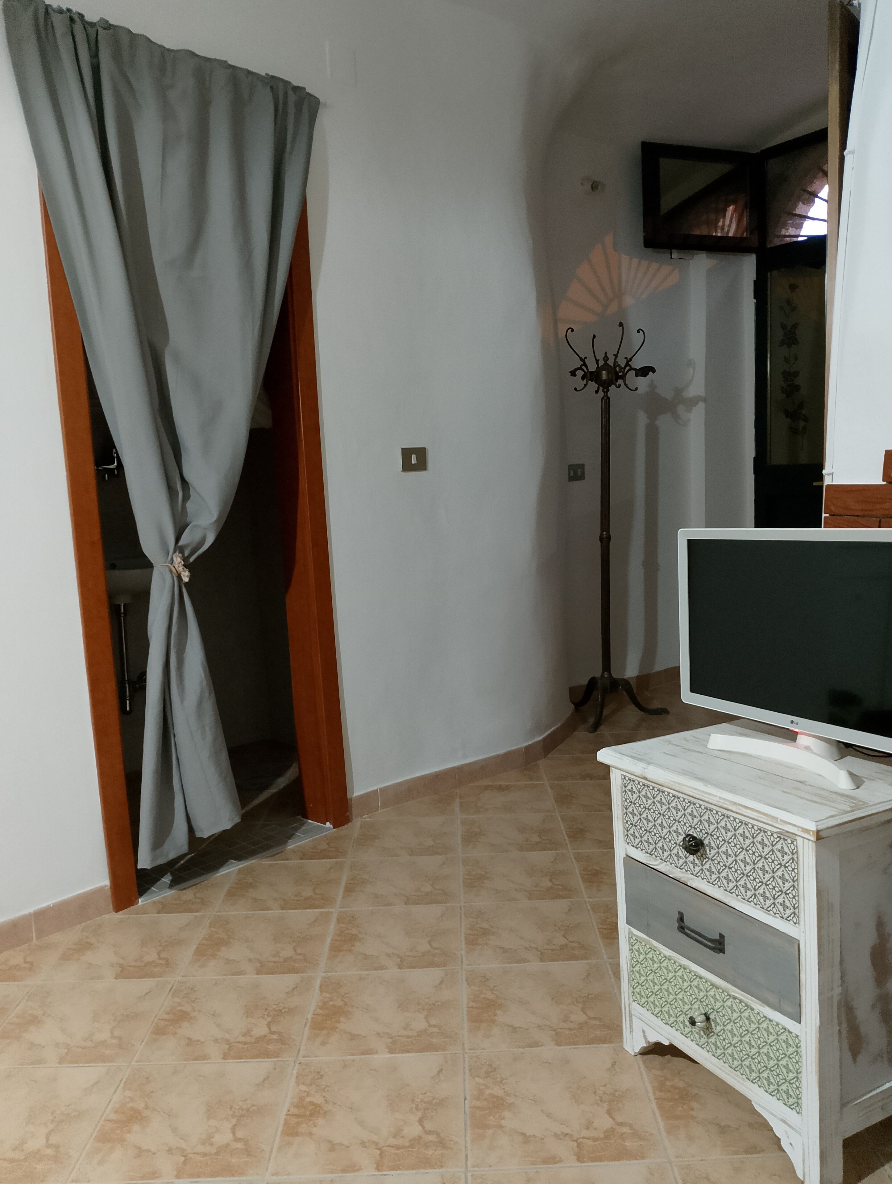 Une pièce avec un téléviseur posé sur un meuble blanc, un lampadaire et un rideau noué en haut d’un cadre de porte.
