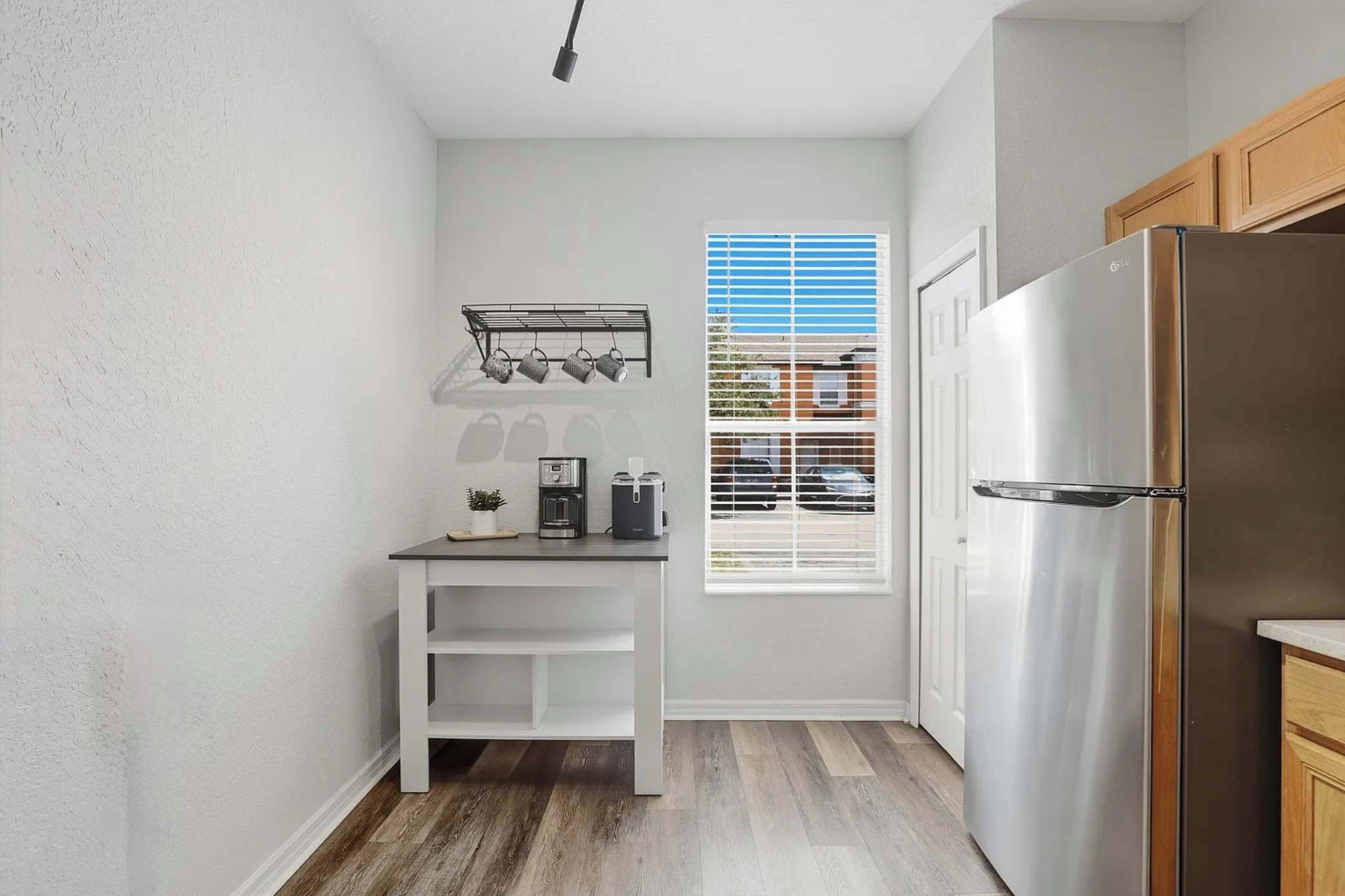 Eine kompakte Küche mit einem Edelstahl-Kühlschrank, einem Fenster mit Jalousien, einem kleinen weißen Tisch mit Kaffeemaschine und einem Metalltrockner.