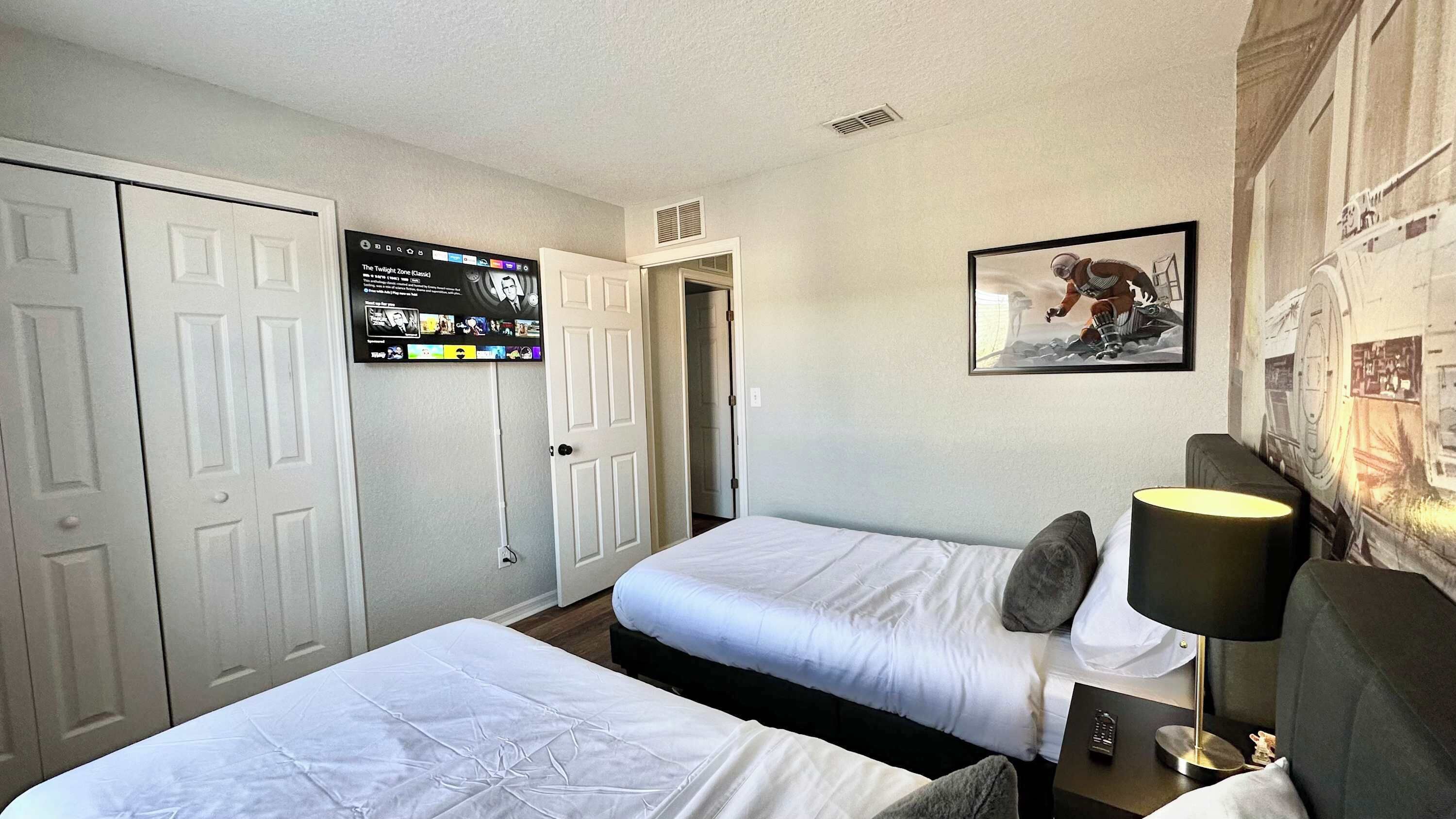 Une chambre d’hôtel avec deux lits simples, un téléviseur fixé au mur diffusant un film, un tableau encadré au mur et une lampe sur une table de chevet.