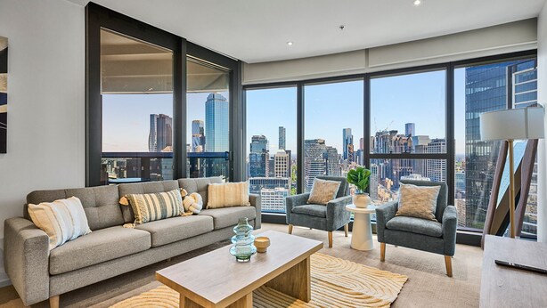 Ein modernes Wohnzimmer mit einem grauen Sofa, Sesseln und einem Couchtisch. Große Fenster bieten Blick auf eine Stadtansicht.