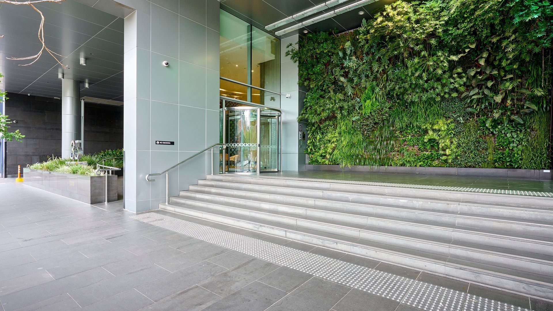 A modern building entrance with a glass elevator, a no smoking sign, and a green vertical garden wall.
