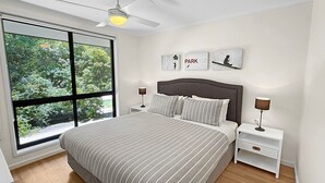 Een slaapkamer met een groot raam, een bed met gestreept beddengoed, twee nachtkastjes met lampen en wanddecoratie.