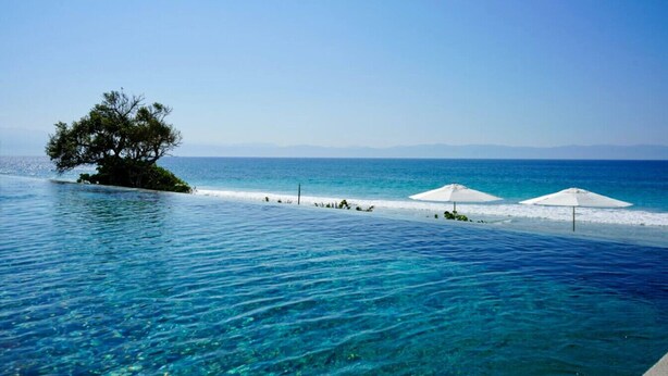 Uma piscina azul límpida com uma árvore numa pequena ilha, dois guarda-sóis brancos e vista para o mar e montanhas ao longe.