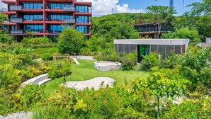 Um edifício moderno com várias varandas e janelas amplas, rodeado por vegetação exuberante e um jardim bem cuidado com um elemento aquático circular.