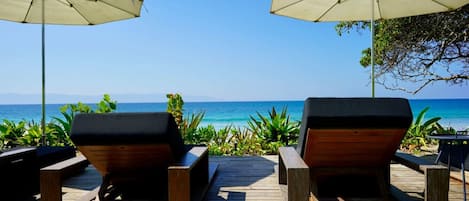 Two wooden lounge chairs with cushions, positioned on a wooden deck with two beige umbrellas providing shade. The deck overlooks a clear blue ocean with a distant shoreline.