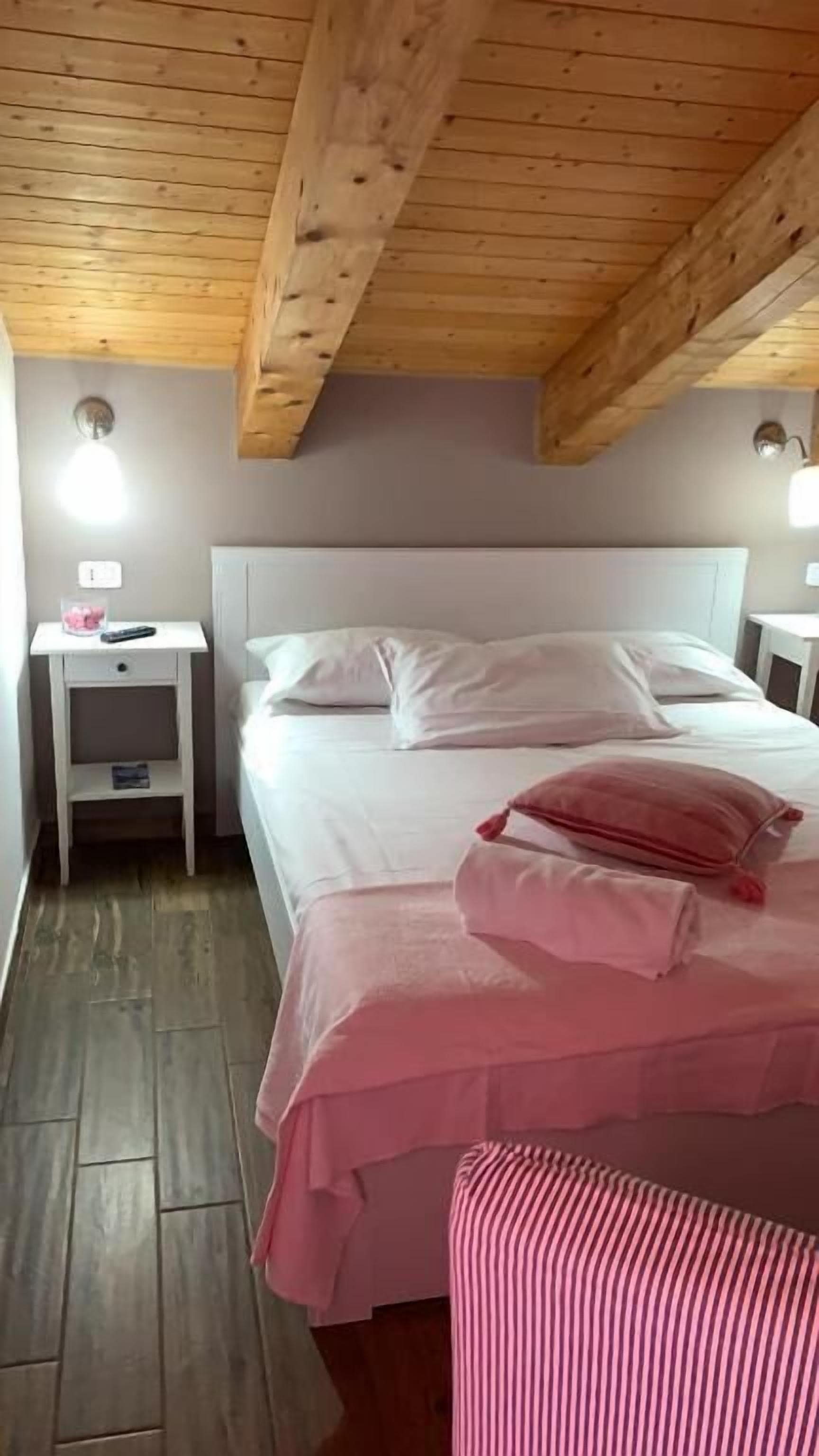 Et soveværelse med træloft, seng med hvidt og pink sengetøj, et natbord med en fjernbetjening og en lille hylde.
