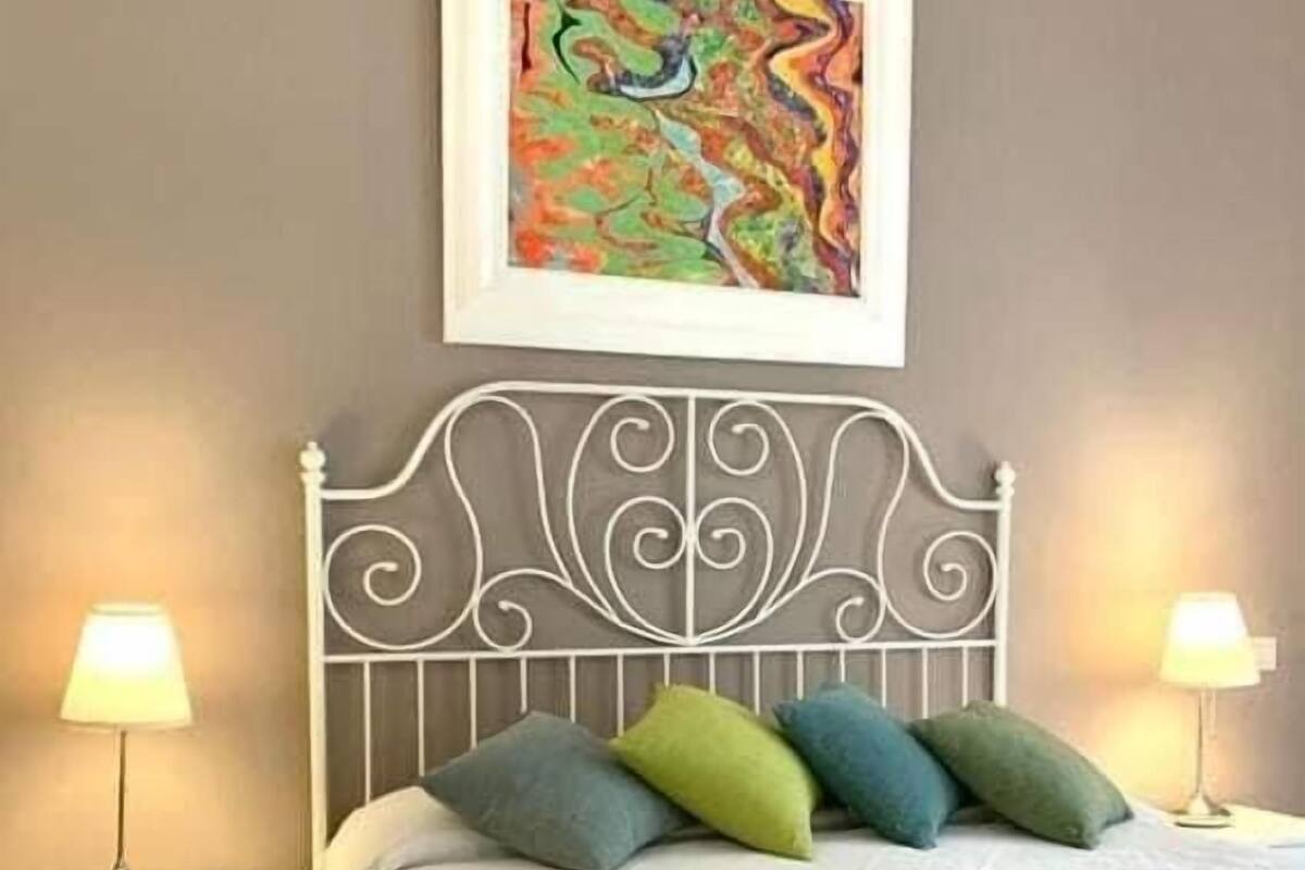 Un lit bien fait, recouvert d’une couvre-lit blanche et agrémenté de coussins décoratifs, avec une tête de lit en métal, des lampes de chevet et un tableau abstrait encadré accroché au mur.