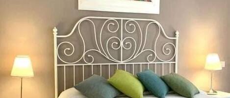 A neatly made bed with a white bedspread and decorative pillows, a metal headboard, bedside lamps, and a framed abstract painting on the wall.