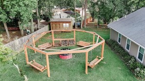 Une terrasse circulaire en bois, dotée de bancs, d’un foyer et d’une petite structure en bois, située dans un jardin.