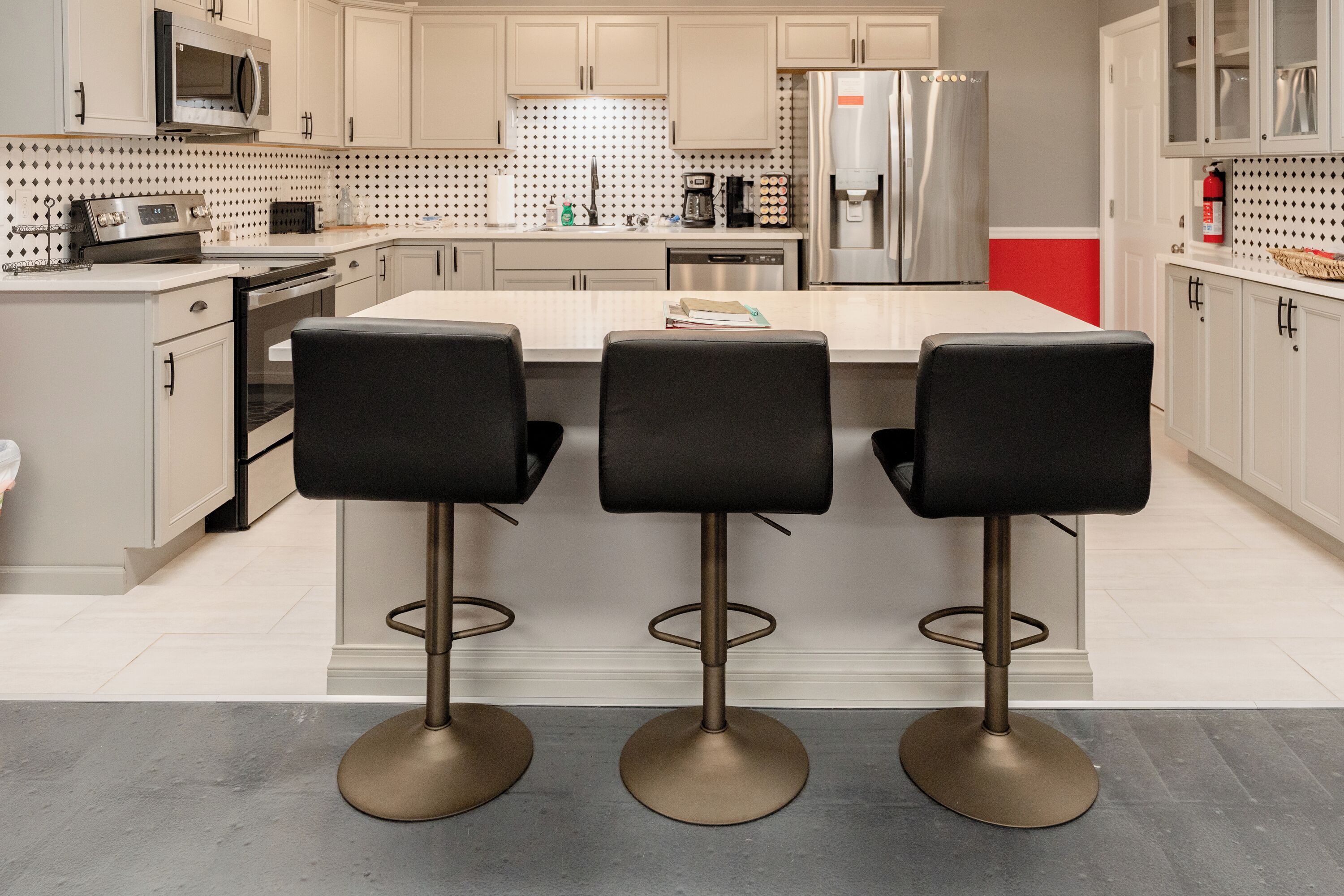 A modern kitchen with three black bar stools, stainless steel appliances, and a tiled backsplash.