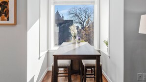 Zona de menjador amb taula i cadires, finestra que mostra un edifici i arbres, i un quadre al mur.
