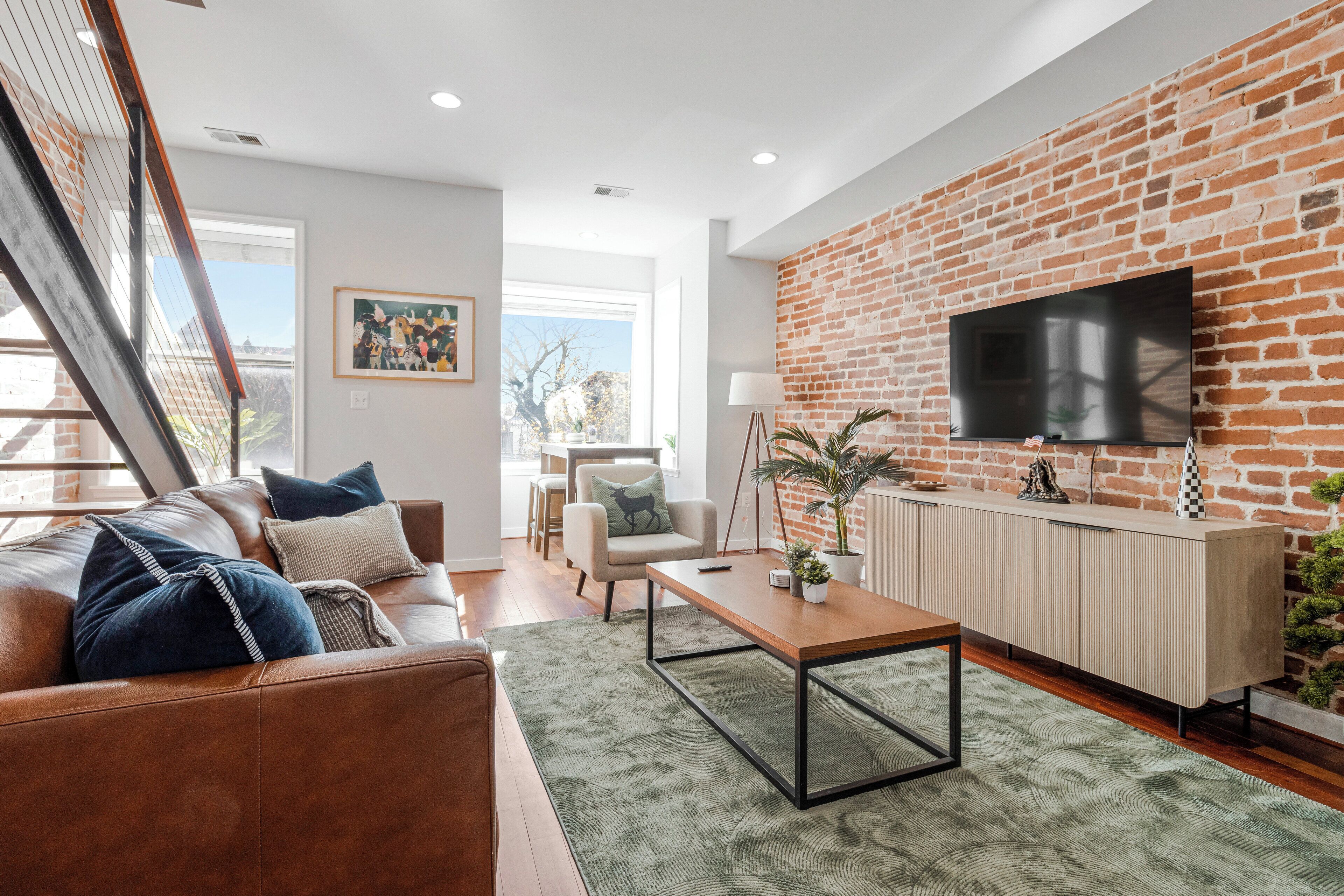 Un salon moderne avec un mur en briques, un téléviseur à écran plat, une table basse en bois, un canapé en cuir et un coin repas comprenant une chaise et une table.