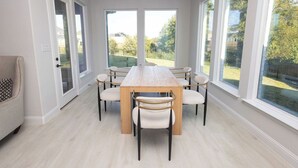 Ein Essbereich mit einem Holztisch und sechs Stühlen, großen Fenstern mit Gartenblick und einem hellen Boden.