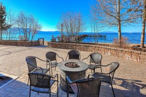 Een terras buiten met een vuurkuil en stoelen, met uitzicht op een waterpartij met een steiger en bergen op de achtergrond.