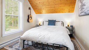 A bedroom with a wooden ceiling, a bed with white bedding, a guitar, and a window with blinds.