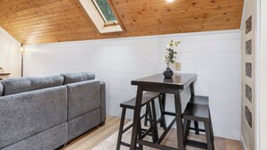 A cozy room with a wooden ceiling, a grey sofa, a small table with a vase, and a window.