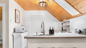 A modern kitchen with a wooden ceiling, white cabinetry, a central island with stools, and a microwave on a cart.