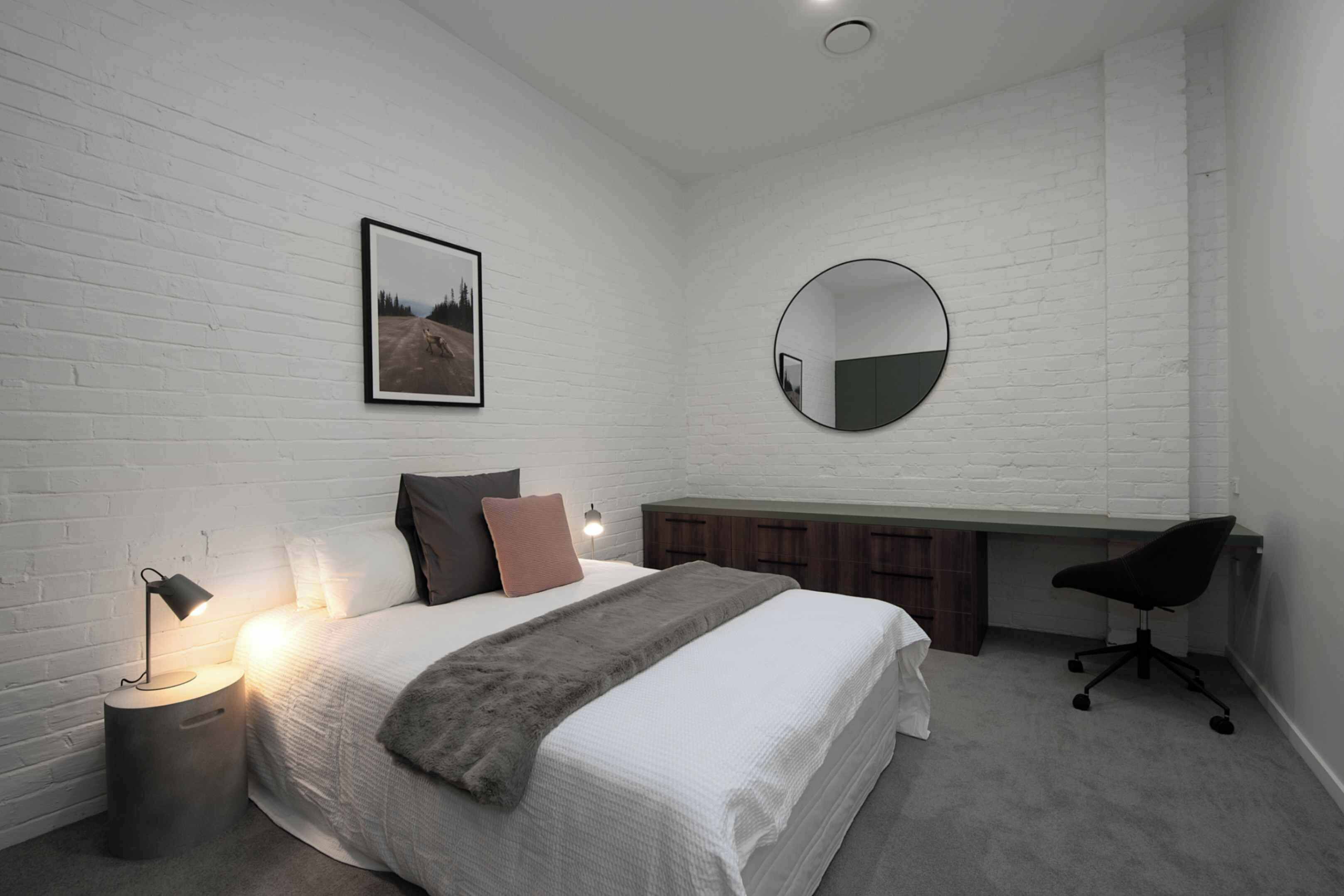 A bedroom with a bed, bedside lamp, a round mirror, a desk, and a chair.