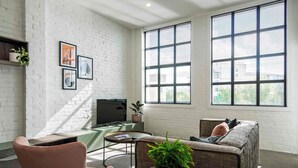 A modern living room with a white brick wall, large windows, a flat-screen TV, a sofa, a chair, and a small table with a plant. - Wow! Luxury inner-city two bedroom apartment (Dunedin)