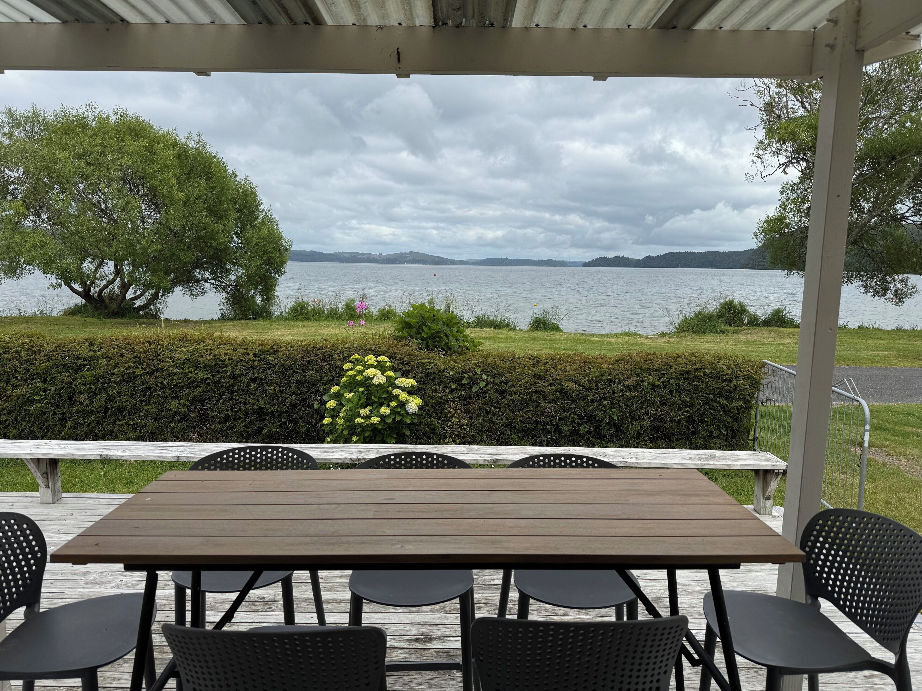 Ein überdachter Außenbereich mit Holztisch und schwarzen Stühlen, mit Blick auf einen See und eine entfernte Uferlinie.
